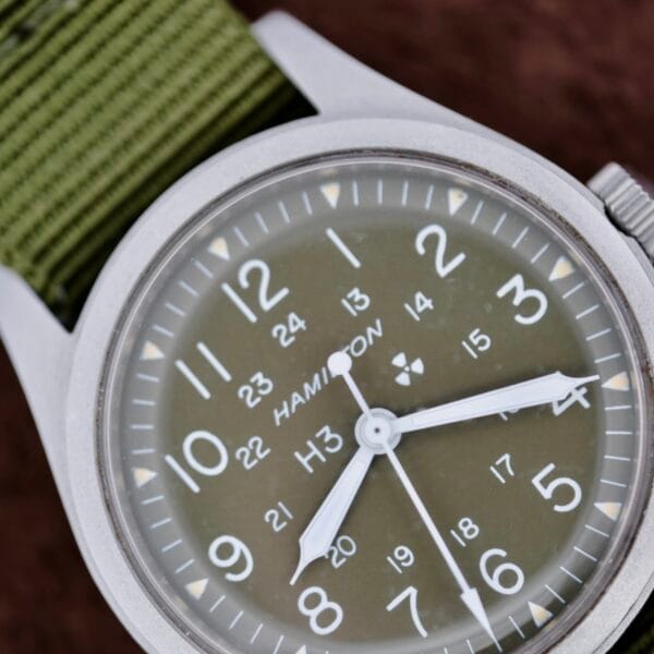 Close-up of the Hamilton Khaki H3 Reference 9219 wristwatch with an olive green face and silver casing, featuring white Arabic numerals for both 12-hour and 24-hour time, a white hour and minute hand, a red-tipped second hand, and a green fabric strap.