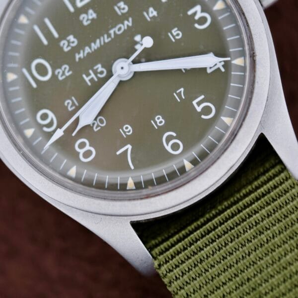 Close-up of a metallic Hamilton Khaki H3 Reference 9219 wristwatch with a green dial, white numerals, both 12 and 24-hour markings, white hands, and a green fabric strap.