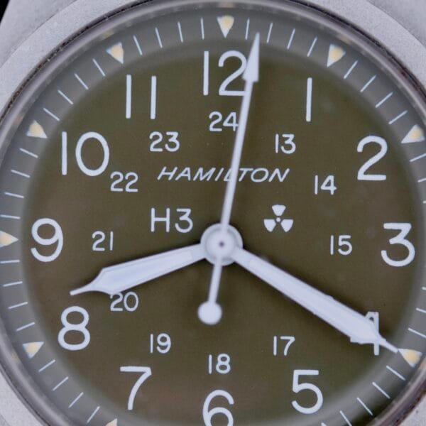 A close-up of the Hamilton Khaki H3 Reference 9219 watch features white hour and minute hands, numerical markings in 12-hour and 24-hour formats, a small nuclear symbol near the center, set against an olive green background.