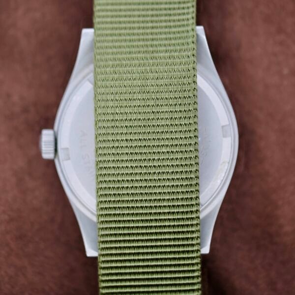 The Hamilton Khaki H3 Reference 9219 with a light green fabric strap lies face down on a brown surface, revealing its watch case partially hidden by the central strap position.
