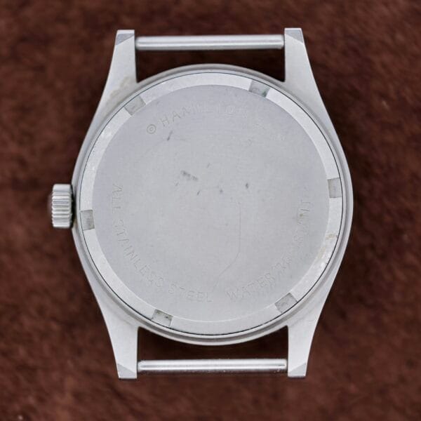 The image displays the back of a Hamilton Khaki H3 Reference 9219 stainless steel wristwatch case, strap unattached. ALL STAINLESS STEEL and WATER RESISTANT are etched on the back beneath the brand name. The background is a textured brown surface.