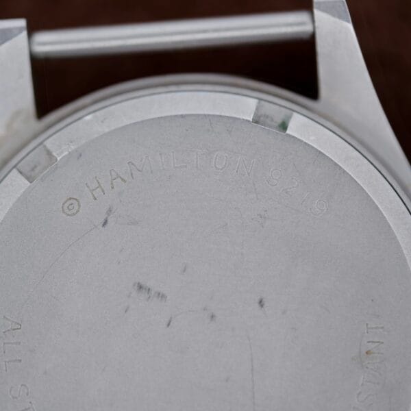 Close-up of the Hamilton Khaki H3 Reference 9219 watch back, engraved with © Hamilton 9219 and showcasing a brushed texture with visible tool marks. A watch strap holder appears at the images top.