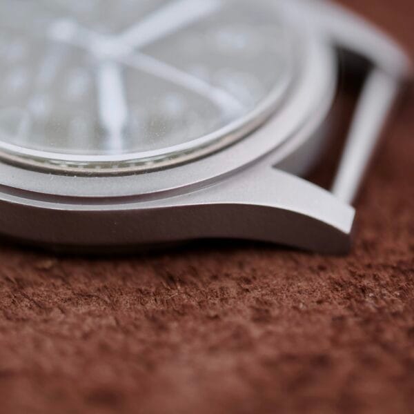 Close-up image of the Hamilton Khaki H3 Reference 9219s silver case, strapless. The blurred watch face reveals minimal details on a textured brown surface, emphasizing its smooth metallic edges and sleek design.