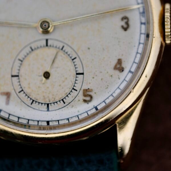 This close-up showcases a timeless 1940s Vacheron Constantin Caltrava V453 wristwatch with a vintage gold case. The white watch face bears charming aging spots, thin black hands, a small secondary dial, and partially visible numerals 3-5. Elegantly attached to a dark strap.