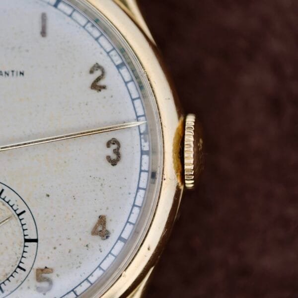 Close-up of a 1940s Vacheron Constantin Caltrava V453 with its timeless face displaying numbers 1-3, a speckled white background, gold-colored casing, right-side crown, and inner rim minute markers.