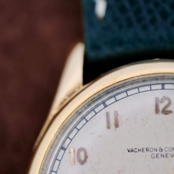 A close-up of the 1940s Vacheron Constantin Caltrava V453 wristwatch reveals its worn white dial and faded hour markers. The gold case edge peeks through next to a dark textured strap, featuring the text Vacheron & Co and Geneve, epitomizing timeless elegance.