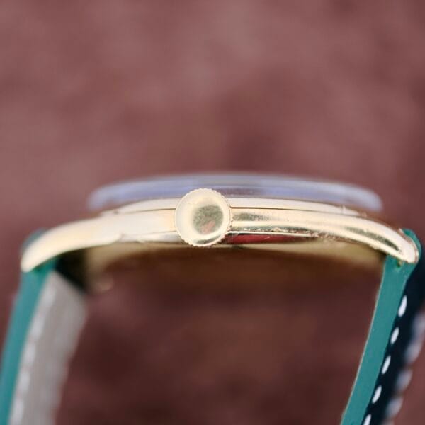 This image showcases a vintage 1940s Vacheron Constantin Caltrava V453 watch. It features a gold case with a ridged crown and a dark green strap accented by light stitching. The soft-focus brown background enhances its timeless appeal.
