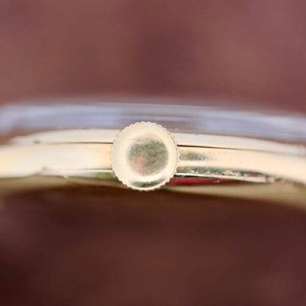 Close-up of a vintage 1940s Vacheron Constantin Caltrava V453 wristwatchs side, highlighting the round, ridged crown for time adjustment. The blurred background accentuates the watchs shiny metallic surface.
