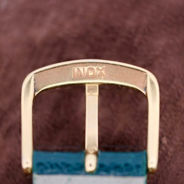A close-up of a gold-toned metal belt buckle engraved with INOX, attached to a blue and grey strap, set against a brown background, reminiscent of the 1940s Vacheron Constantin Caltrava V453 style.
