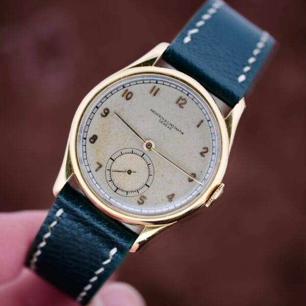 The 1940s Vacheron Constantin Caltrava V453 showcases a close-up of its vintage wristwatch with a beige dial, gold casing, and dark leather strap. This timeless piece includes Arabic numerals, minute markings, and a small seconds subdial at the 6 oclock position.