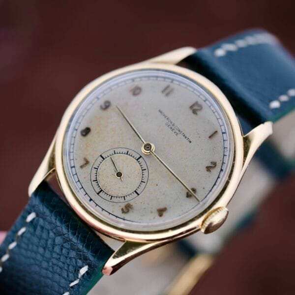 A close-up of the 1940s Vacheron Constantin Caltrava V453 features a round, tarnished beige dial encased in gold. Arabic numerals and a seconds subdial add charm, while the dark textured leather strap with white stitching completes its timeless allure.