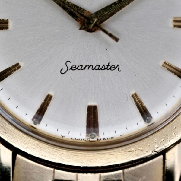 A vintage close-up of the Omega Seamaster L6298 Dauphine Hands watch reveals a white dial with gold hour markers and hands, exuding timeless elegance. Seamaster is scripted on the dial, and T SWISS MADE T appears at the bottom, alongside a glimpse of its graceful gold case.