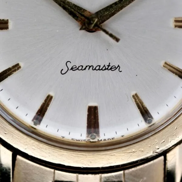 A vintage close-up of the Omega Seamaster L6298 Dauphine Hands watch reveals a white dial with gold hour markers and hands, exuding timeless elegance. Seamaster is scripted on the dial, and T SWISS MADE T appears at the bottom, alongside a glimpse of its graceful gold case.