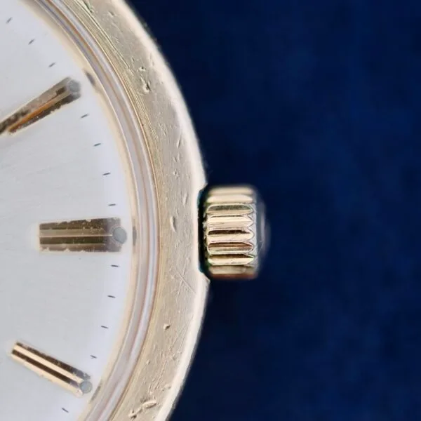 A close-up of the Omega Seamaster L6298 Dauphine Hands showcases a vintage watch face with a silver finish and visible texture. The fluted crown contrasts against a deep blue backdrop, complemented by partially seen hour markers, enhancing its classic charm.