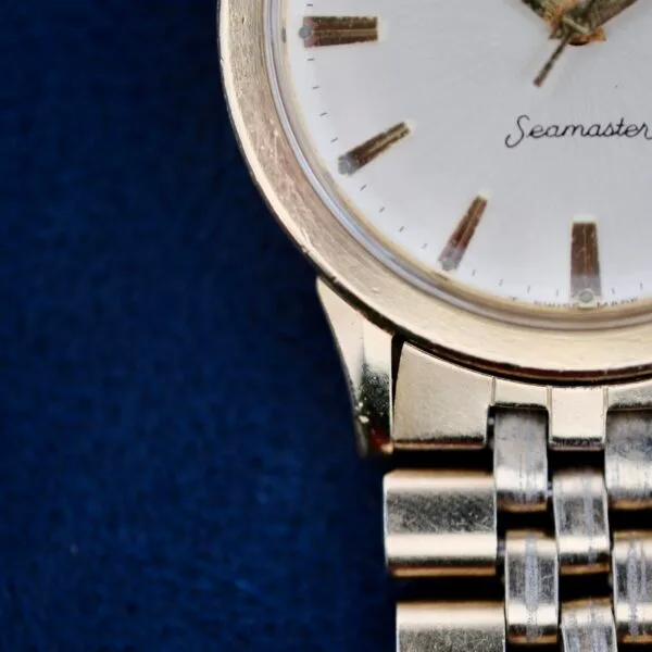 Close-up of an Omega Seamaster L6298 Dauphine Hands watch against a dark blue background, featuring a white face with gold hour markers and hands. Its metal band is polished with a linked design showcasing timeless elegance.