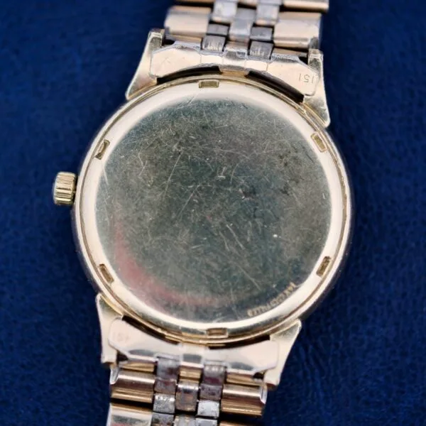 A close-up of the Omega Seamaster L6298 Dauphine Hands showcases its scratched silver-tone case. This vintage wristwatch features a metal link band with alternating gold and silver sections, resting against textured dark blue fabric to enhance its timeless charm.