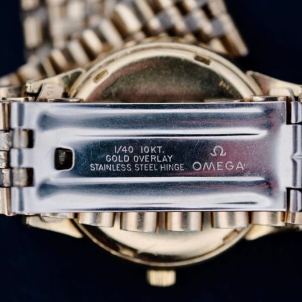 A close-up image captures the vintage allure of an Omega Seamaster L6298 Dauphine Hands wristwatchs metal clasp, featuring 1/40 10KT. GOLD OVERLAY STAINLESS STEEL HINGE OMEGA, emphasizing its metallic charm and gold finish.
