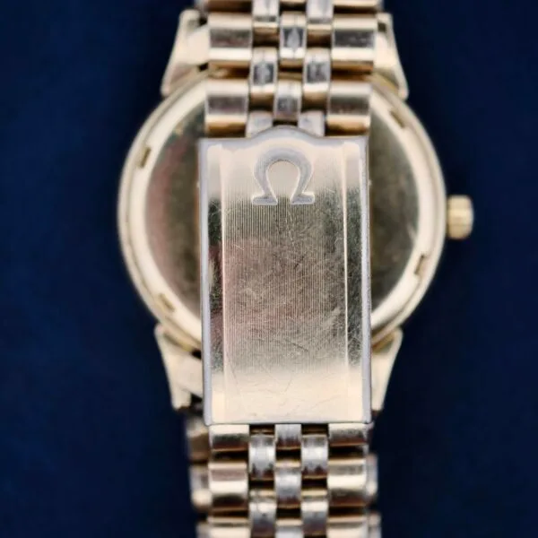 Close-up of the back of a vintage Omega Seamaster L6298 Dauphine Hands wristwatch with a metal link strap. The clasp features an engraved Omega logo, showing some scratches and wear. A dark blue background enhances its timeless appeal.