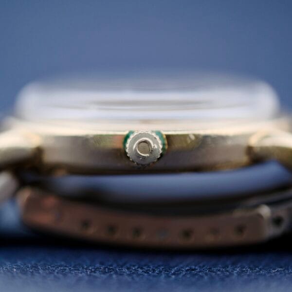 Side view of the Omega Seamaster L6298 Dauphine Hands watch on a blue background, focusing on the metallic, textured crown. The strap is partially visible below.