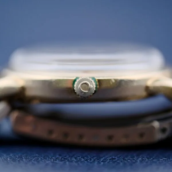 Side view of the Omega Seamaster L6298 Dauphine Hands watch on a blue background, focusing on the metallic, textured crown. The strap is partially visible below.