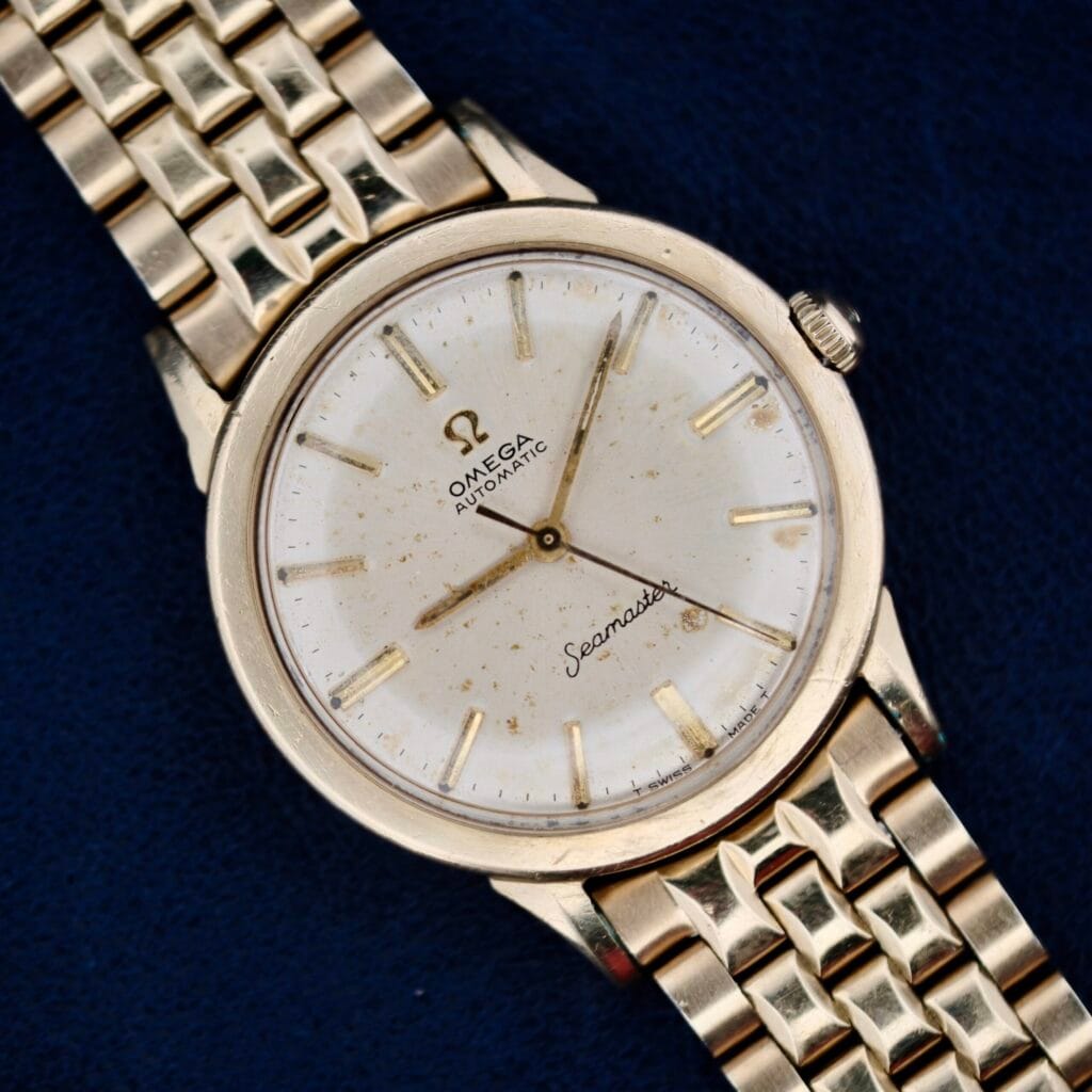 The Omega Seamaster L6298 Automatic wristwatch with a textured gold band and round face is showcased on a dark blue surface. This vintage timepiece features a white dial, gold hour markers, two hands, and a small crown on the right side.