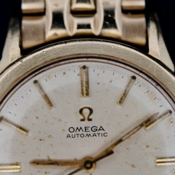 A close-up of the Omega Seamaster L6298 Automatic wristwatch reveals a gold-toned metal band and a weathered white dial with simple gold hour markers and hands. The iconic Omega logo and AUTOMATIC are prominently displayed below 12 oclock, showcasing its timeless allure.