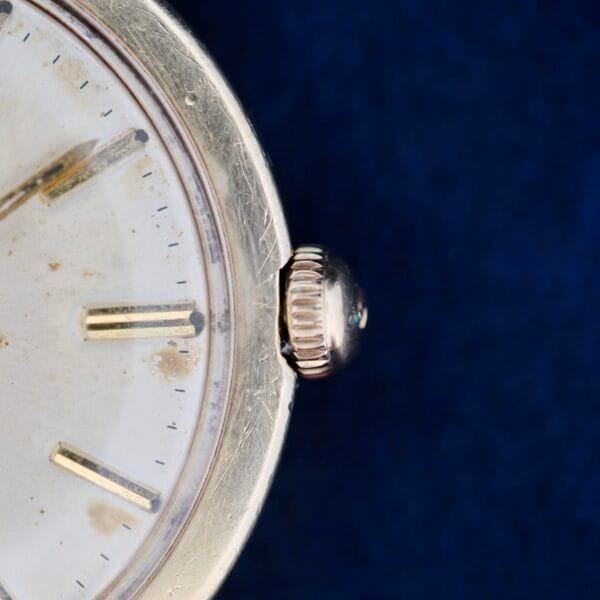 A close-up of the Omega Seamaster L6298 Automatic watch showcases its round metallic case and textured crown. The watch face, with minute and hour hands and simple hour markers, exudes timeless elegance against a rich dark blue fabric backdrop that enhances its classic charm.