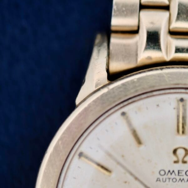 A close-up focuses on the Omega Seamaster L6298 Automatic wristwatch, showcasing its vintage gold face and brand name. Partial markings and hands are visible, while the classic gold strap gleams softly against a dark backdrop.
