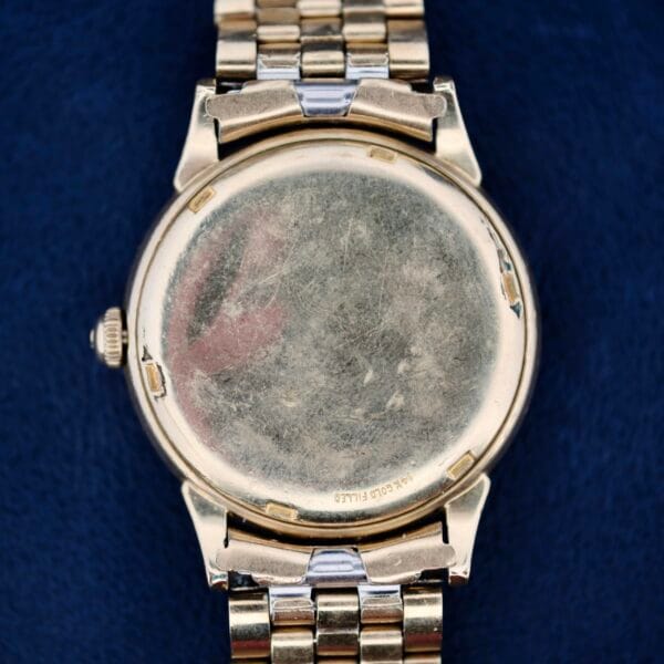 A close-up of the back of an Omega Seamaster L6298 Automatic, featuring a vintage, tarnished gold finish and a metal link band. The round case has a small crown, with engraved text visible against a textured dark blue background.