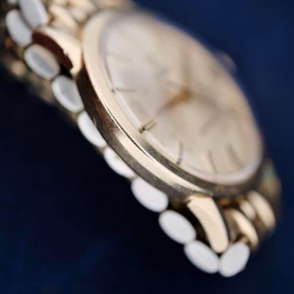 A close-up of the Omega Seamaster L6298 Automatic wristwatch showcases its gold-toned case and bracelet against a dark blue background. The blurred watch face with indistinct time markings and hands emphasizes the bracelets rounded links.