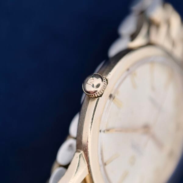 The Omega Seamaster L6298 Automatic showcases its gold-toned case and crown, highlighting a partially visible white dial with hour markings. Set against a dark blue backdrop, the scene reveals the classic charm of its metal bracelet.