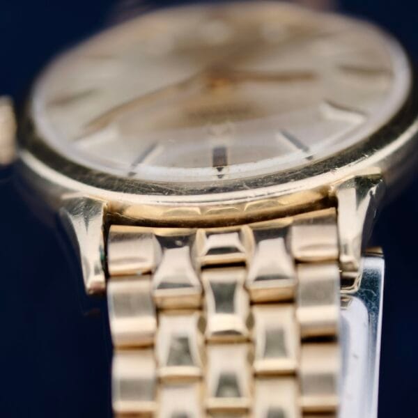 Close-up of the Omega Seamaster L6298 Automatic, highlighting its watch face with slender hour markers and smooth bezel. The metal band displays a detailed interlocking design, while soft lighting enhances its timeless gold tones.