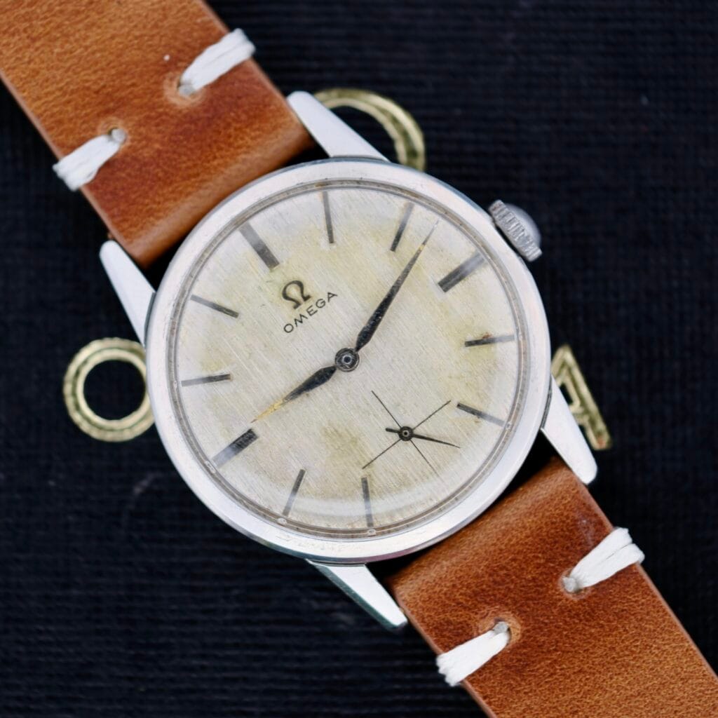 The Omega Ref 14713 Bark Dial wristwatch features a worn beige dial with black hour markers, a seconds subdial, silver-toned hands, and the iconic Omega logo below the 12 oclock marker. A brown leather strap with white stitching enhances its timeless elegance.