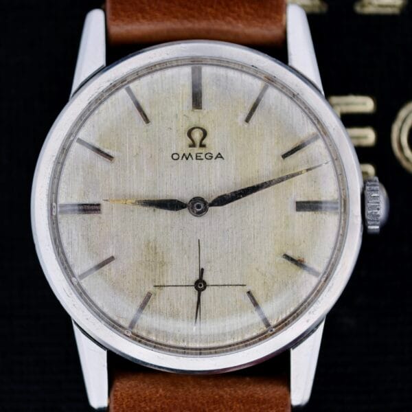 The Omega Ref 14713 Bark Dial wristwatch features a silver case, an aged dial with black hour and minute hands, a small sub-dial at the bottom, and pairs perfectly with a brown leather strap. The Omega logo and name are elegantly placed under the 12 oclock marker.
