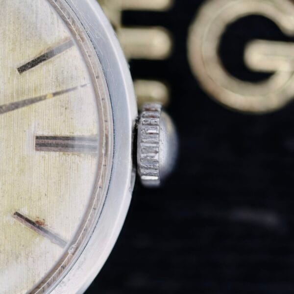 Close-up of an Omega Ref 14713 Bark Dial watch face showing the glass, a silver crown, and part of the dial with visible hour markers. The blurred background features partially obscured golden lettering.