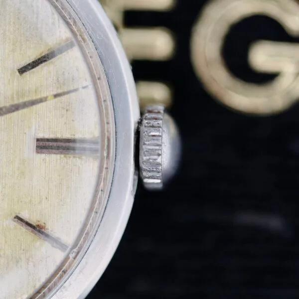 Close-up of an Omega Ref 14713 Bark Dial watch face showing the glass, a silver crown, and part of the dial with visible hour markers. The blurred background features partially obscured golden lettering.