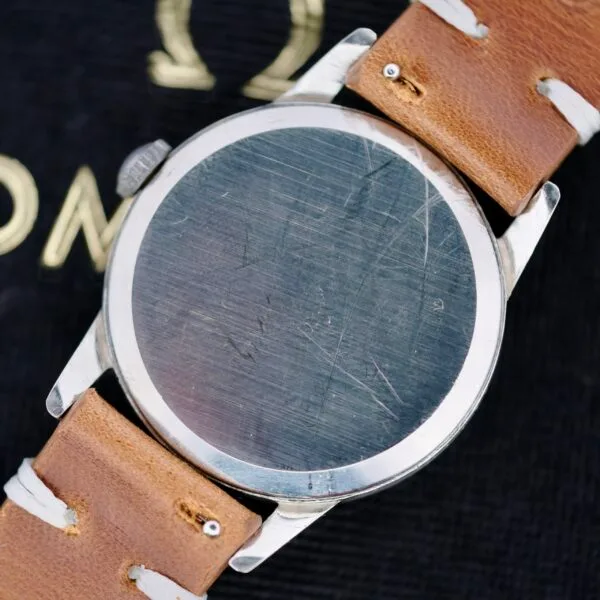 A close-up of the Omega Ref 14713 Bark Dial wristwatch displays a scratched metal back and brown leather strap, set against a background featuring part of an Omega logo in gold on black.