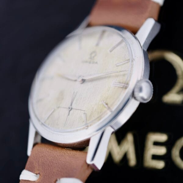 The Omega Ref 14713 Bark Dial wristwatch features a light face, slender metal hands, and slim hour markers within a metallic case. A brown leather strap enhances its classic appeal, while a blurred embossed OMEGA logo in the dark background amplifies its timeless charm.