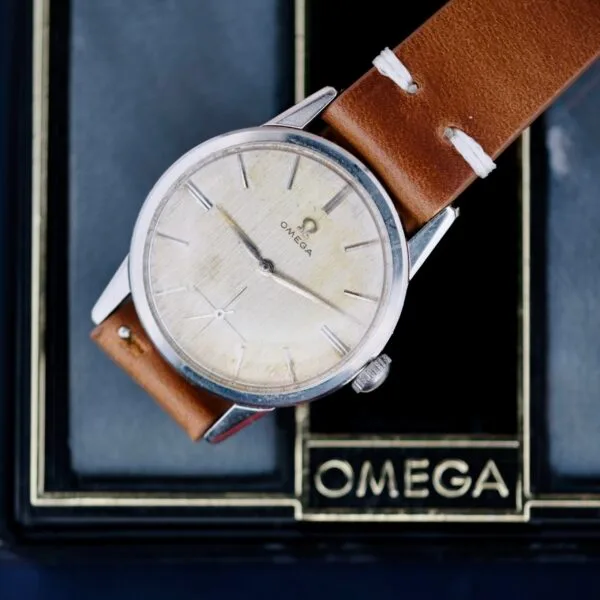 The Omega Ref 14713 Bark Dial is a classic wristwatch with a vintage charm, featuring a silver-tone round face, minimalist tick marks, and a brown leather strap. It is elegantly displayed in a case adorned with the Omega logo in gold lettering.