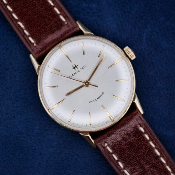 The 1960s Hamilton Automatic Sunburst Dial features a vintage design with a white dial, gold hour markers, and hands. It is paired with a dark brown leather strap with white stitching and beautifully displayed on a dark blue background.