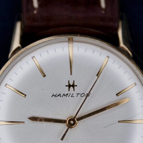 Close-up of a vintage 1960s Hamilton Automatic Sunburst Dial wristwatch with a brown leather strap. The silver watch face showcases minimalistic gold hour markers and hands. Hamilton is elegantly displayed below the twelve oclock marker, enhancing its timeless charm.