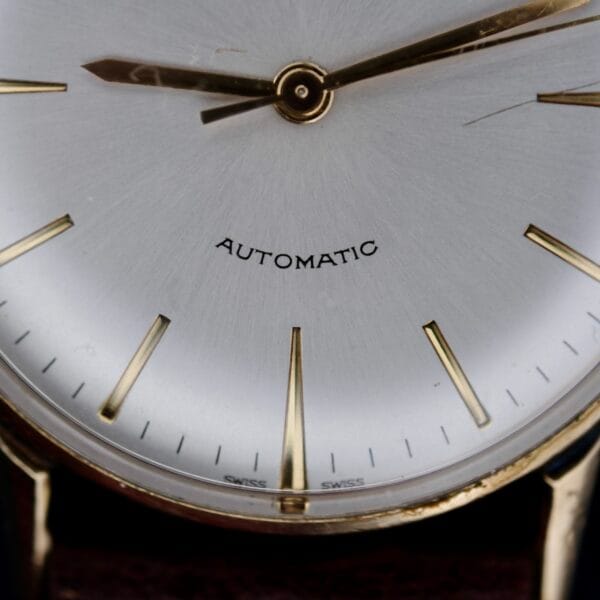Close-up of a 1960s Hamilton Automatic Sunburst Dial watch with a silver face, gold hour markers, and two gold hands. AUTOMATIC is printed above the center, capturing timeless elegance against a dark background.