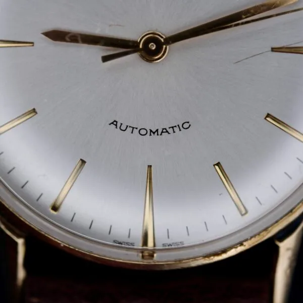 Close-up of a 1960s Hamilton Automatic Sunburst Dial watch with a silver face, gold hour markers, and two gold hands. AUTOMATIC is printed above the center, capturing timeless elegance against a dark background.