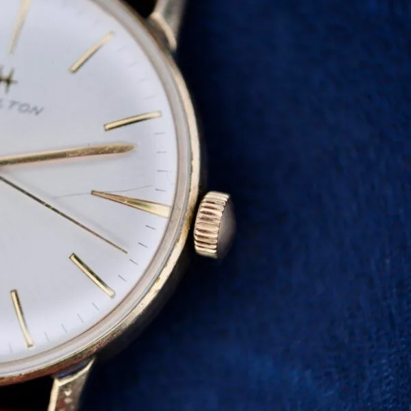 This close-up features a 1960s Hamilton Automatic Sunburst Dial with a white face, gold hour markers and hands. Encased in gold with a ridged crown on its right, its set against deep blue fabric, and partially obscured text enhances the mystery of this elegant timepiece.