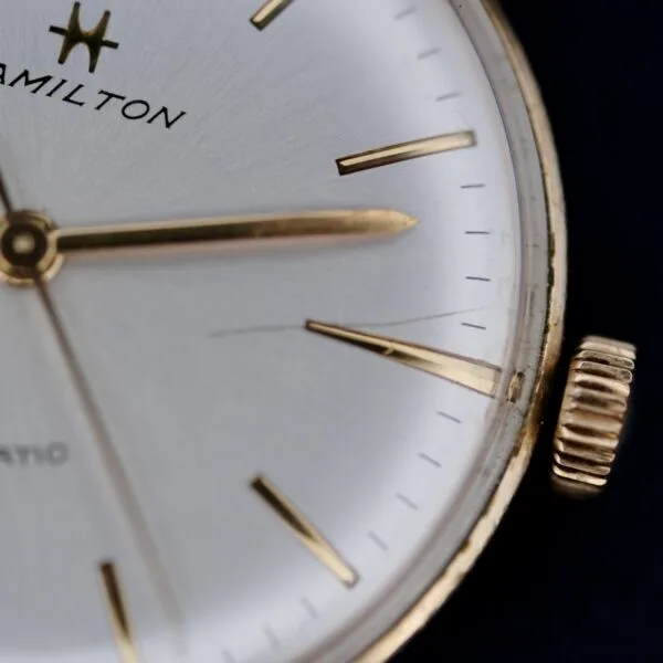 Close-up of a 1960s Hamilton Automatic Sunburst Dial watch with a silver face, thin gold hour markers and hands, a gold casing with ridged crown, and the logo plus partial Automatic visible, highlighting its classic automatic movement.