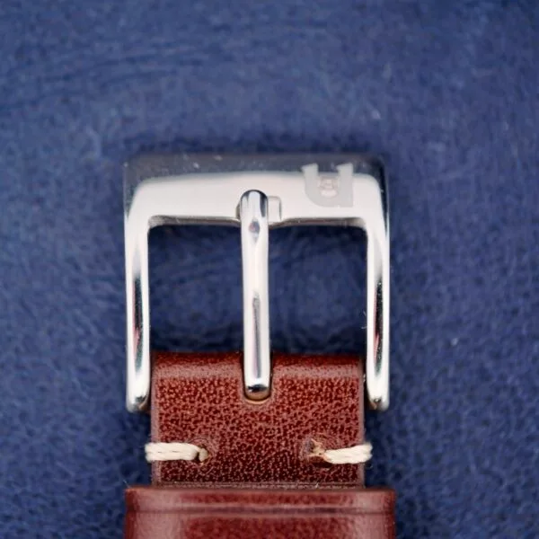 A detailed view of the 1960s Hamilton Automatic Sunburst Dial showcases a shiny silver belt buckle on a vintage brown leather strap with beige stitching, set against a textured blue background.