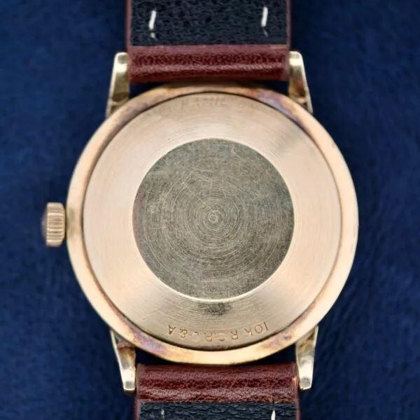 The image shows the back of a 1960s Hamilton Automatic Sunburst Dial wristwatch, featuring a round, gold-toned case and brown leather strap. Its worn, scratched surface adds charm, with 10K R.G.P. engraved on the bottom. The watch lies gracefully on a dark blue surface.