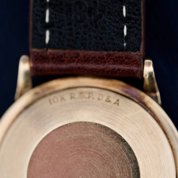 Close-up of the back of a vintage 1960s Hamilton Automatic Sunburst Dial gold watch case with 10K R.G.P. D&A engraved. It has a dark leather strap with visible stitching, set against a blurred dark background enhancing its classic appeal.