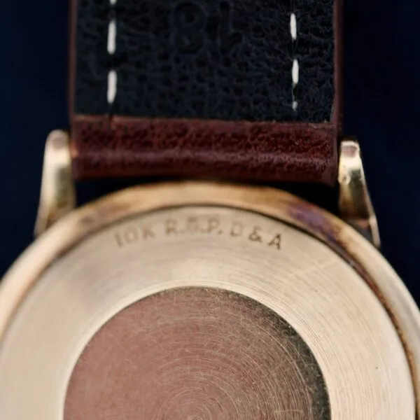 Close-up of the back of a vintage 1960s Hamilton Automatic Sunburst Dial gold watch case with 10K R.G.P. D&A engraved. It has a dark leather strap with visible stitching, set against a blurred dark background enhancing its classic appeal.