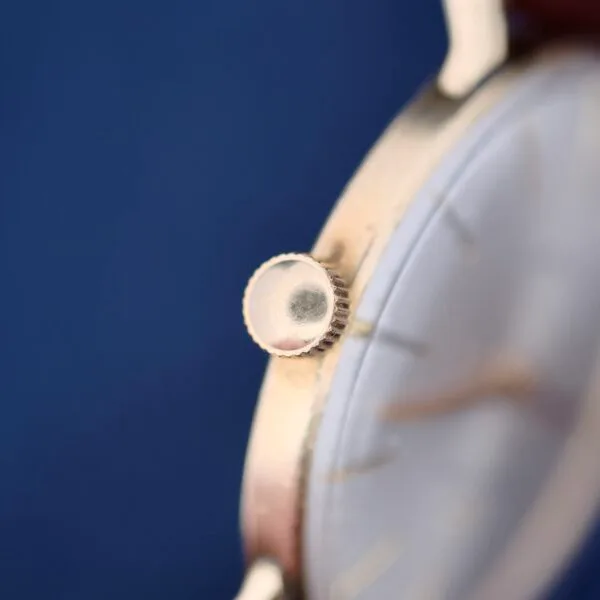 Close-up of the gold ridged crown of a 1960s Hamilton Automatic Sunburst Dial against a blurred blue background. The watch face, with faint hour markers and partially visible elements, hints at its classic analog design.